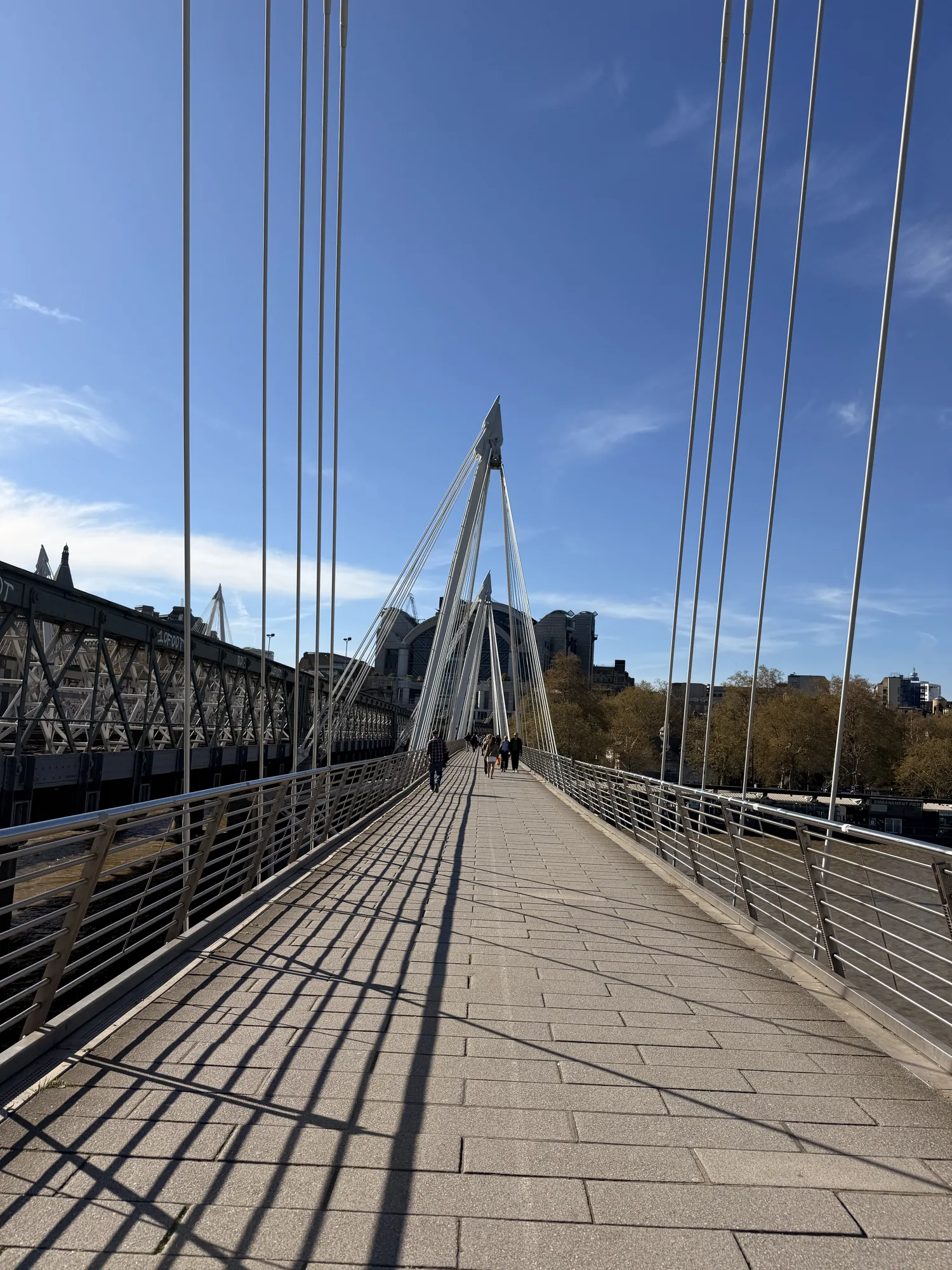 A London footbridge with long railing shadows beneath a blue sky.
