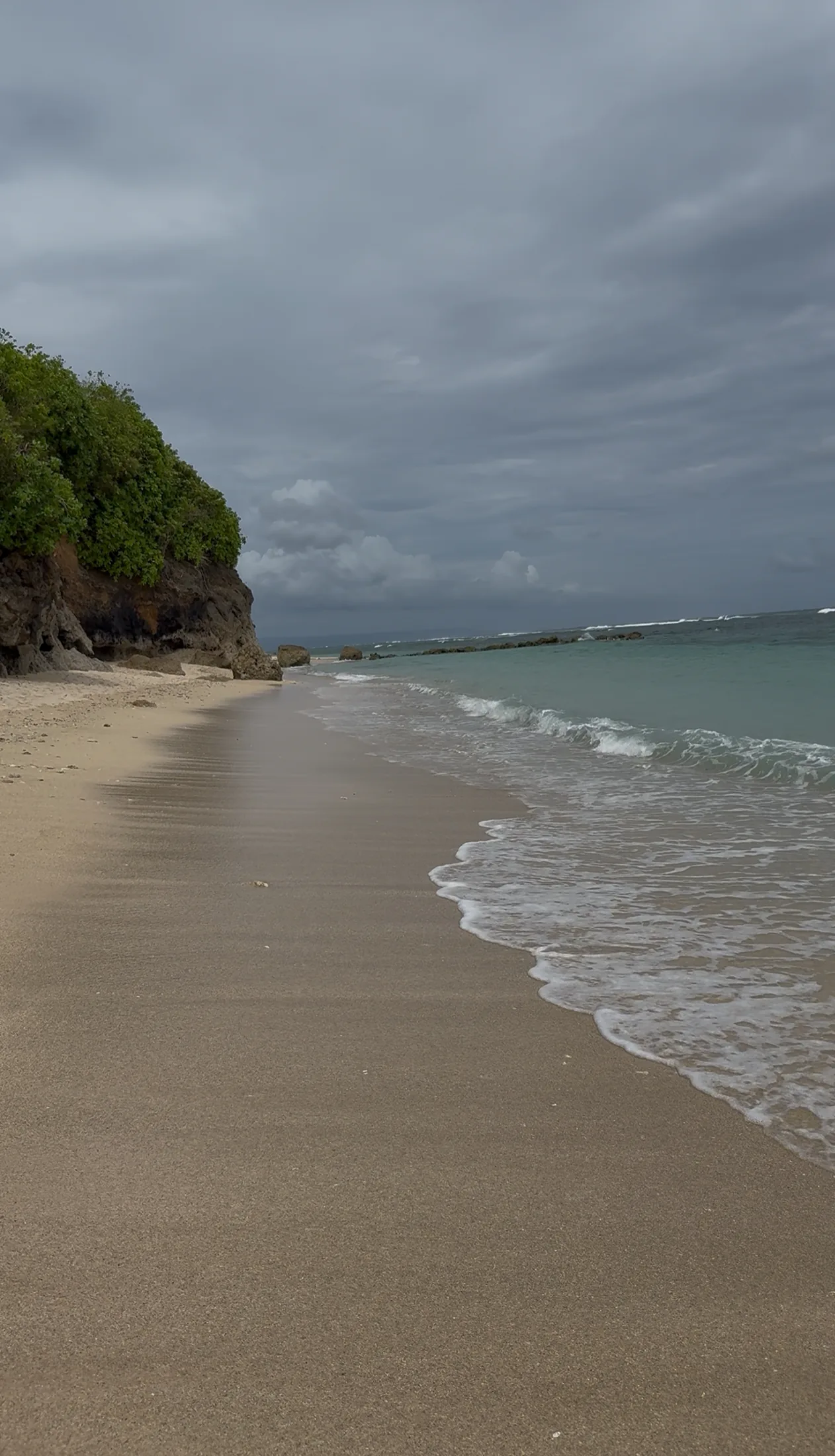 Waves reaching a sandy Bali beach beneath a clouded sky